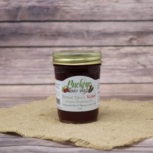 A 9oz glass jar of Buckeye Berry Patch Brother Dave's Kicker Jalapeno Raspberry Jam, on rustic burlap with a wooden background.