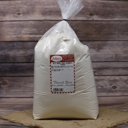 A clear plastic bag of Gold Medal Full Strength Flour, tied with a black twist tie on rustic burlap with a wooden background.